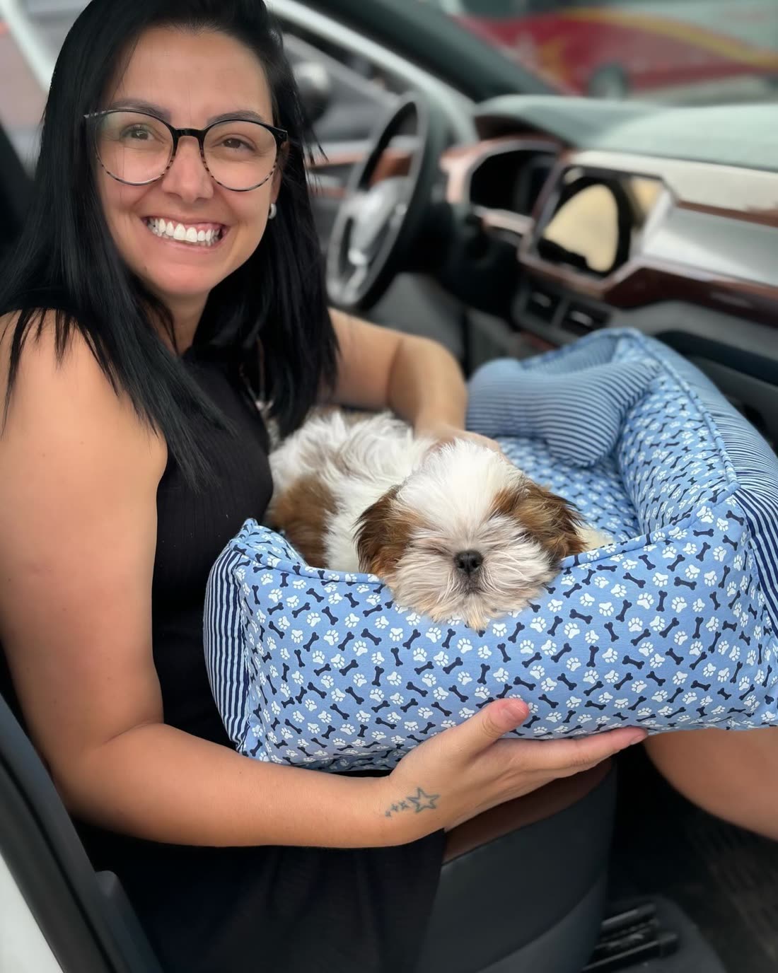 Linda from Tennessee smiling in her car with Sam, a gold and white Shih Tzu puppy, asleep in a blue bed on her lap