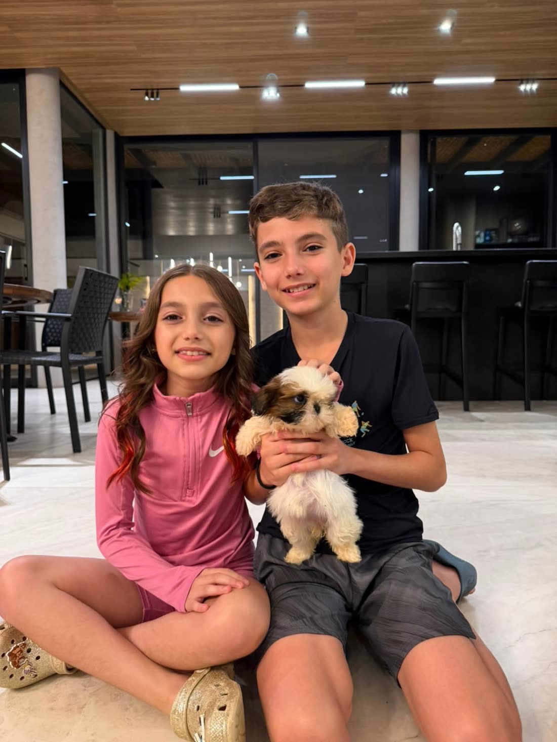 Two children sitting on the floor at home, holding Nate, a brown and white Shih Tzu puppy