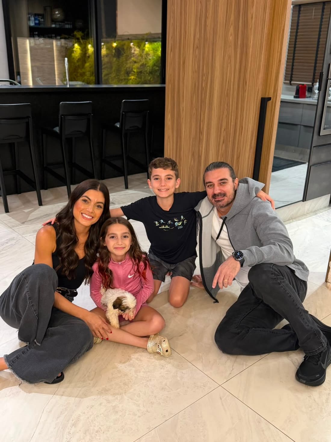 A family of four sitting on their kitchen floor with Nate, a brown and white Shih Tzu puppy, on the daughter’s lap